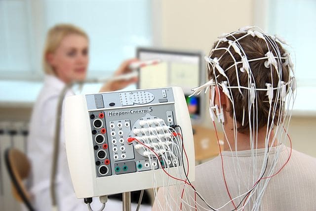 A man with EEG electrodes on his head and female researcher in the background.
